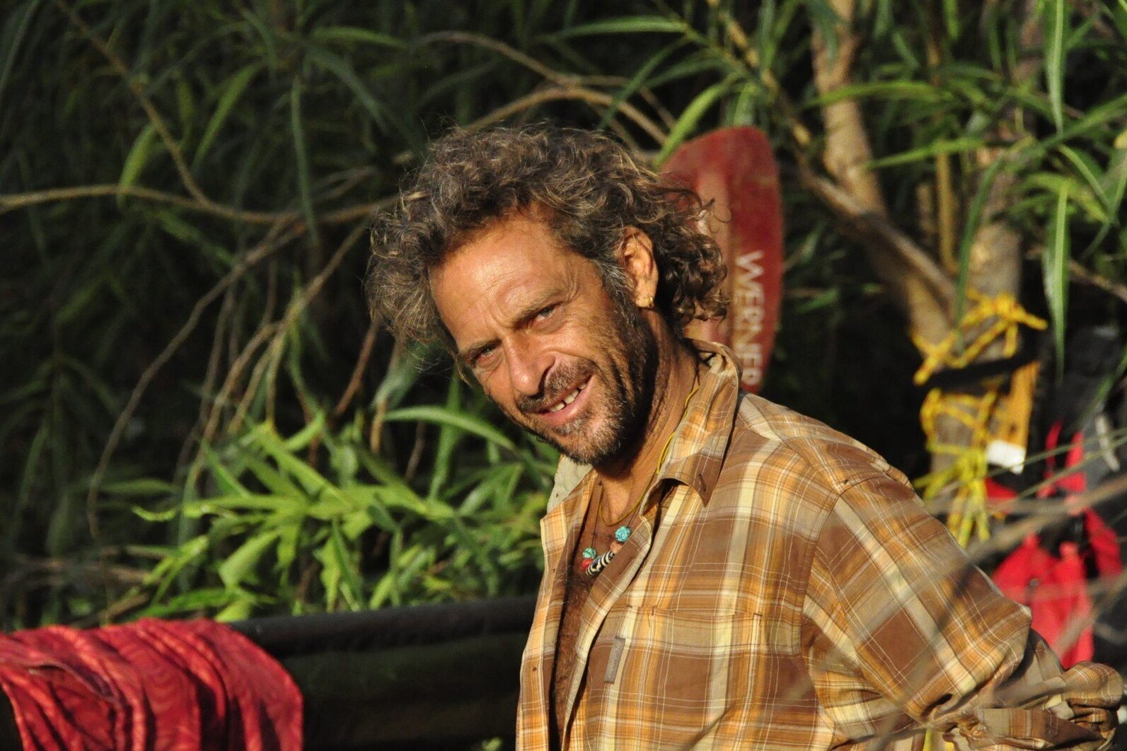 Totem Advenuture A man with curly hair and a checked shirt smiles outdoors, surrounded by the lush greenery of Pré-Saint-Didier, capturing the adventurous spirit of Aosta Valley outdoor experiences. Rafting Kayaking Paragliding Canyoning Aosta Valley