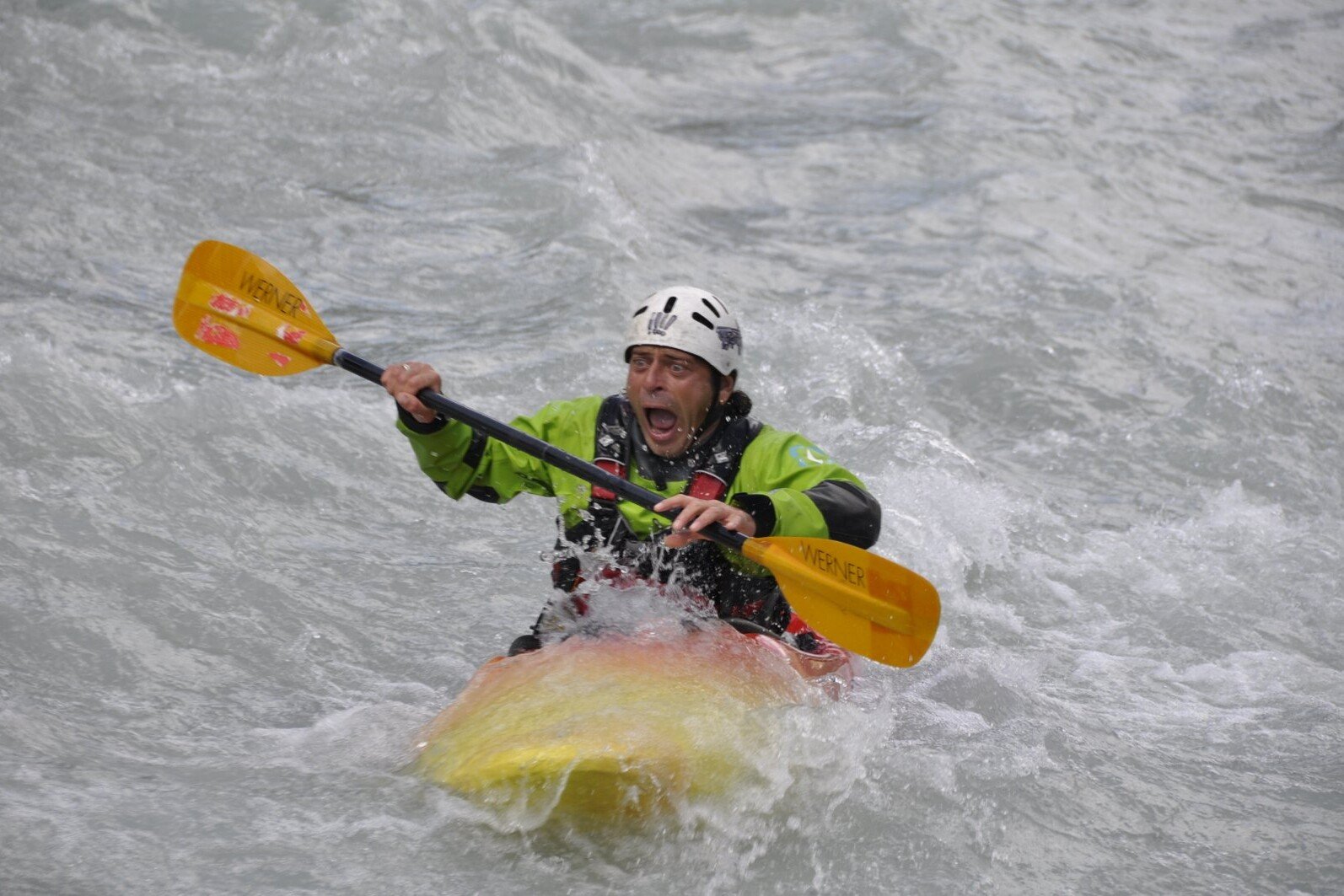 Totem Advenuture Person in green jacket and helmet kayaking through rough waters with a determined expression, enjoying the exciting outdoor experiences in the Aosta Valley. Rafting Kayaking Paragliding Canyoning Aosta Valley