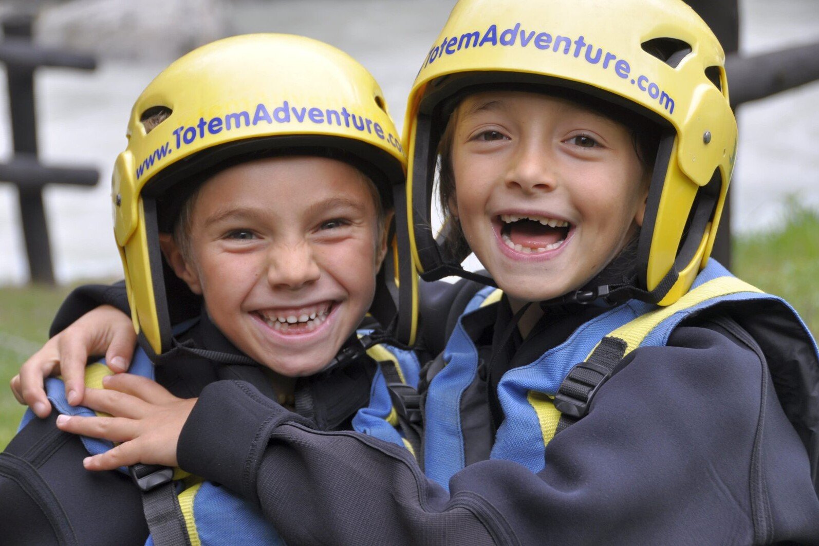 Totem Advenuture Two smiling children in yellow helmets and wetsuits embrace outdoors, ready for an adventure in the beautiful rafting terrain of the Aosta Valley, near Pre Saint Didier. Rafting Kayaking Paragliding Canyoning Aosta Valley