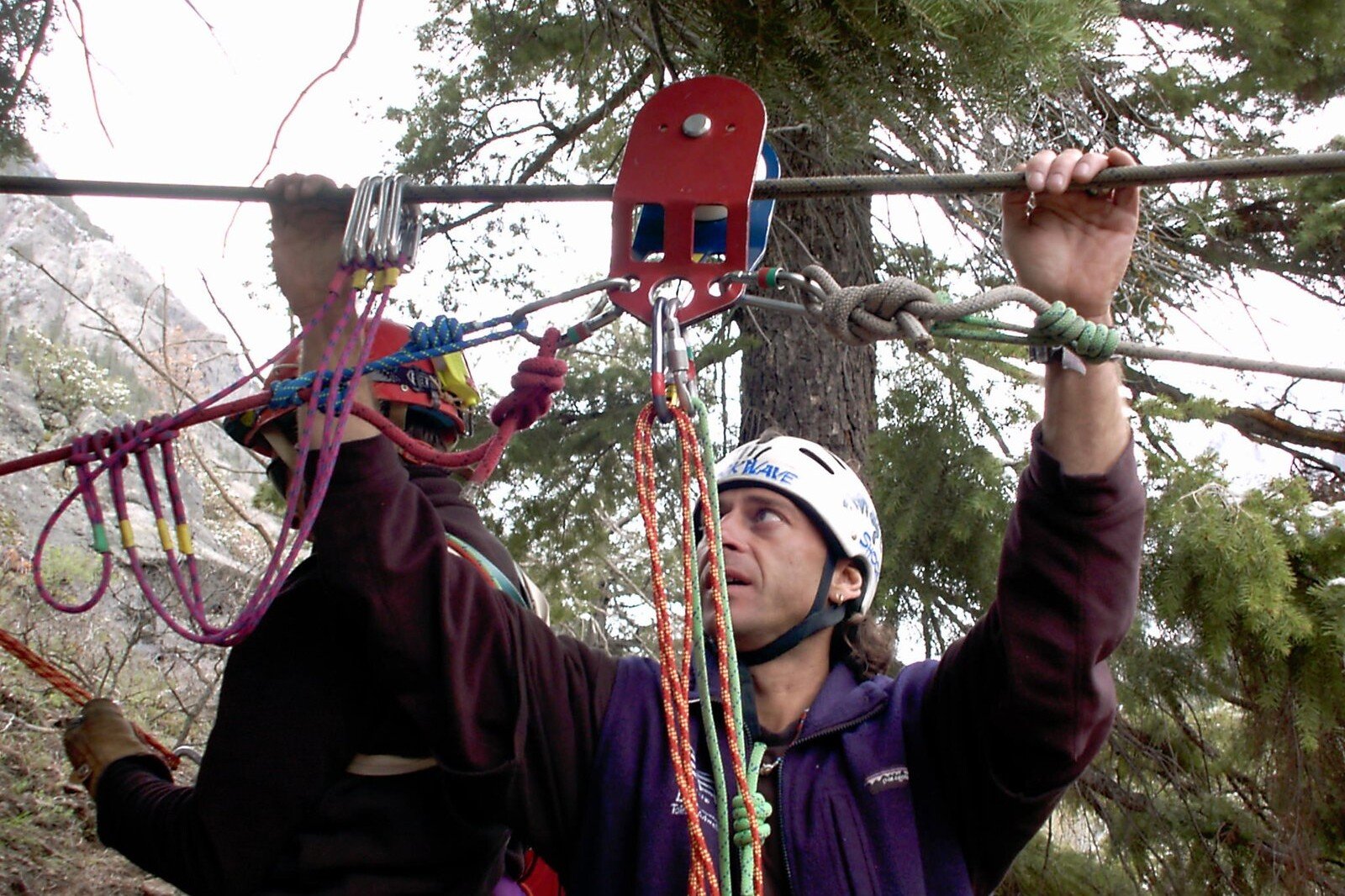 Totem Adventure In a setting among the trees reminiscent of an adventure in the heart of Pré-Saint-Didier, a person wearing a helmet deftly regulates ropes and pulleys, while another is standing nearby. Rafting Kayaking Paragliding Canyoning Aosta Valley