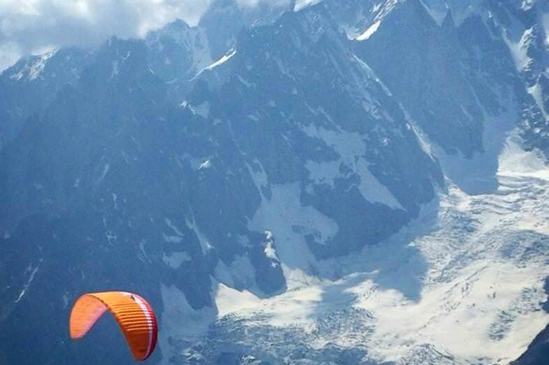 Totem Advenuture A paraglider with an orange and red canopy flies close to snow-capped mountains under partly cloudy skies, embodying the exciting spirit of outdoor experiences in the Aosta Valley. Rafting Kayaking Paragliding Canyoning Aosta Valley