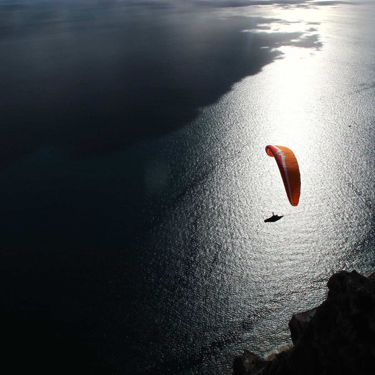 Totem Advenuture A paraglider with an orange canopy flies over the ocean near a rocky coastline, the sunlight reflecting off the water and reminiscent of the exhilarating outdoor experiences in Valle d'Aosta. Rafting Kayaking Paragliding Canyoning Aosta Valley