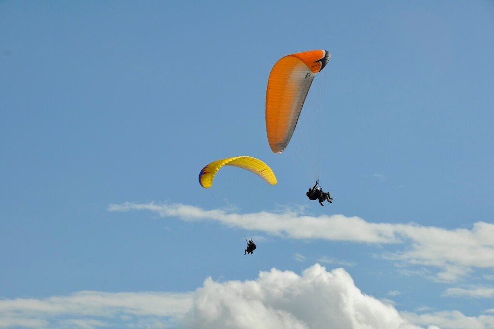 Totem Advenuture Three paragliders with colourful sails soar through a clear blue sky above fluffy white clouds, capturing the spirit of outdoor adventure in the Aosta Valley. Rafting Kayaking Paragliding Canyoning Aosta Valley
