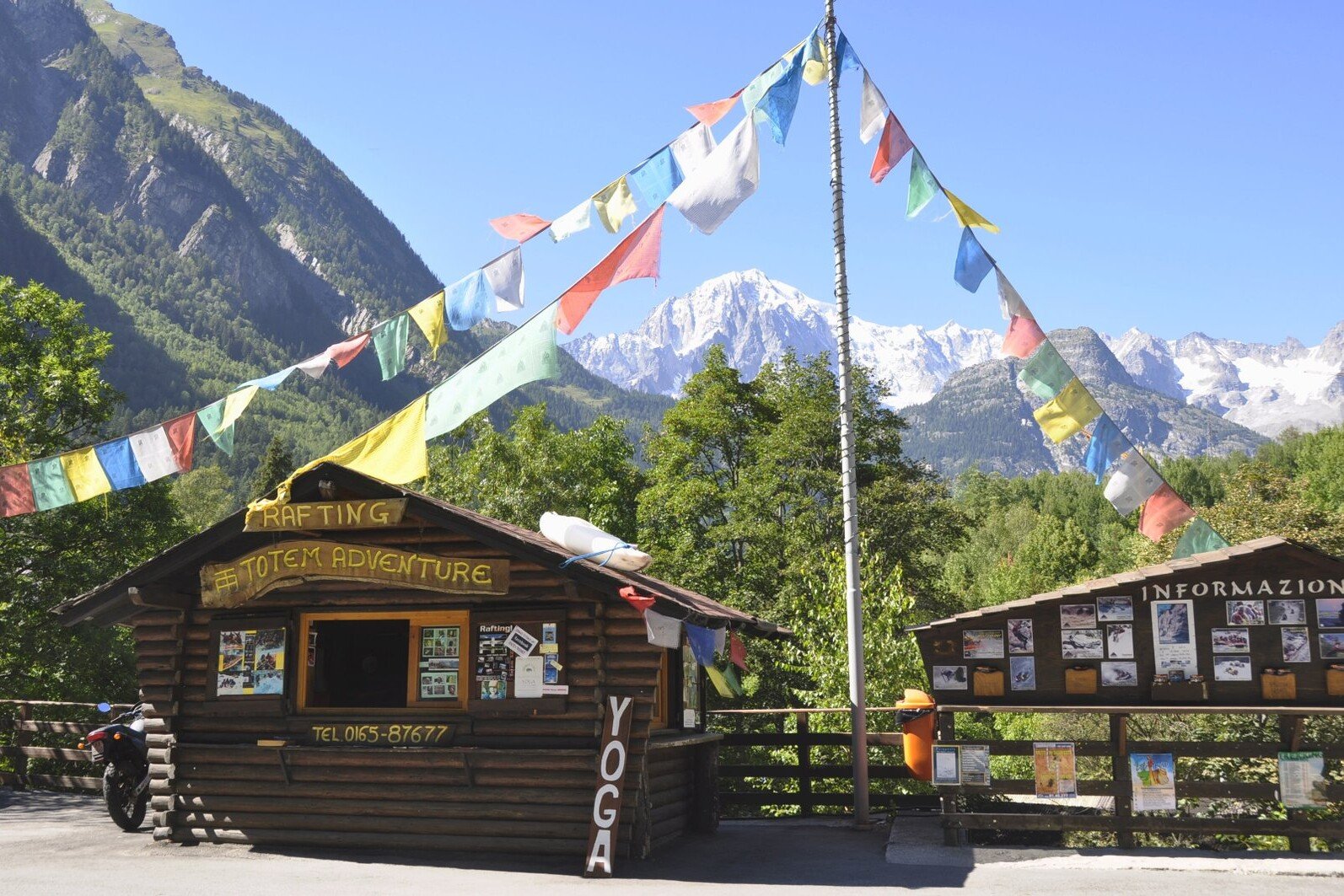 Totem Advenuture Wooden cabin with adventurous signage, colourful flags and a backdrop of mountains. Nearby information panels highlight Aosta Valley kayaking outdoor experiences, inviting thrill-seekers to explore the beautiful landscapes. Rafting Kayaking Paragliding Canyoning Aosta Valley