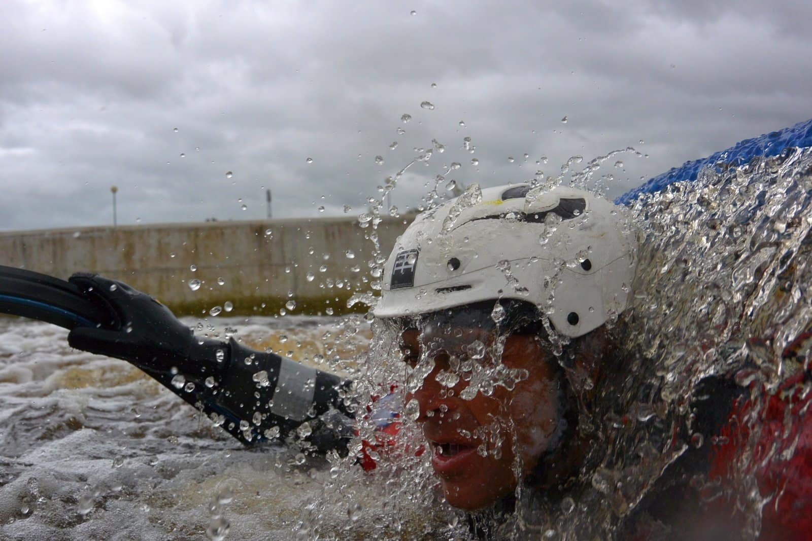 Totem Advenuture A person wearing a white helmet and a wetsuit is submerged in rough water, stretching out amidst splashes, against the backdrop of a cloudy sky. This scene captures the thrill of adventure in the Aosta Valley, where rafting in its dynamic waters offers unparalleled outdoor experiences. Rafting Kayaking Paragliding Canyoning Aosta Valley