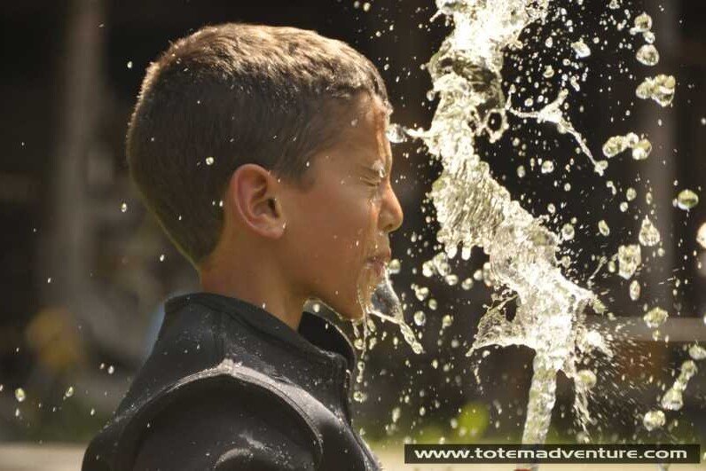 Totem Advenuture A child with his eyes closed immersed in splashing water, wearing a dark jacket and the drops illuminated by the sunlight: a moment of pure adventure reminiscent of experiences in the open-air paradise of the Aosta Valley. Rafting Kayaking Paragliding Canyoning Aosta Valley
