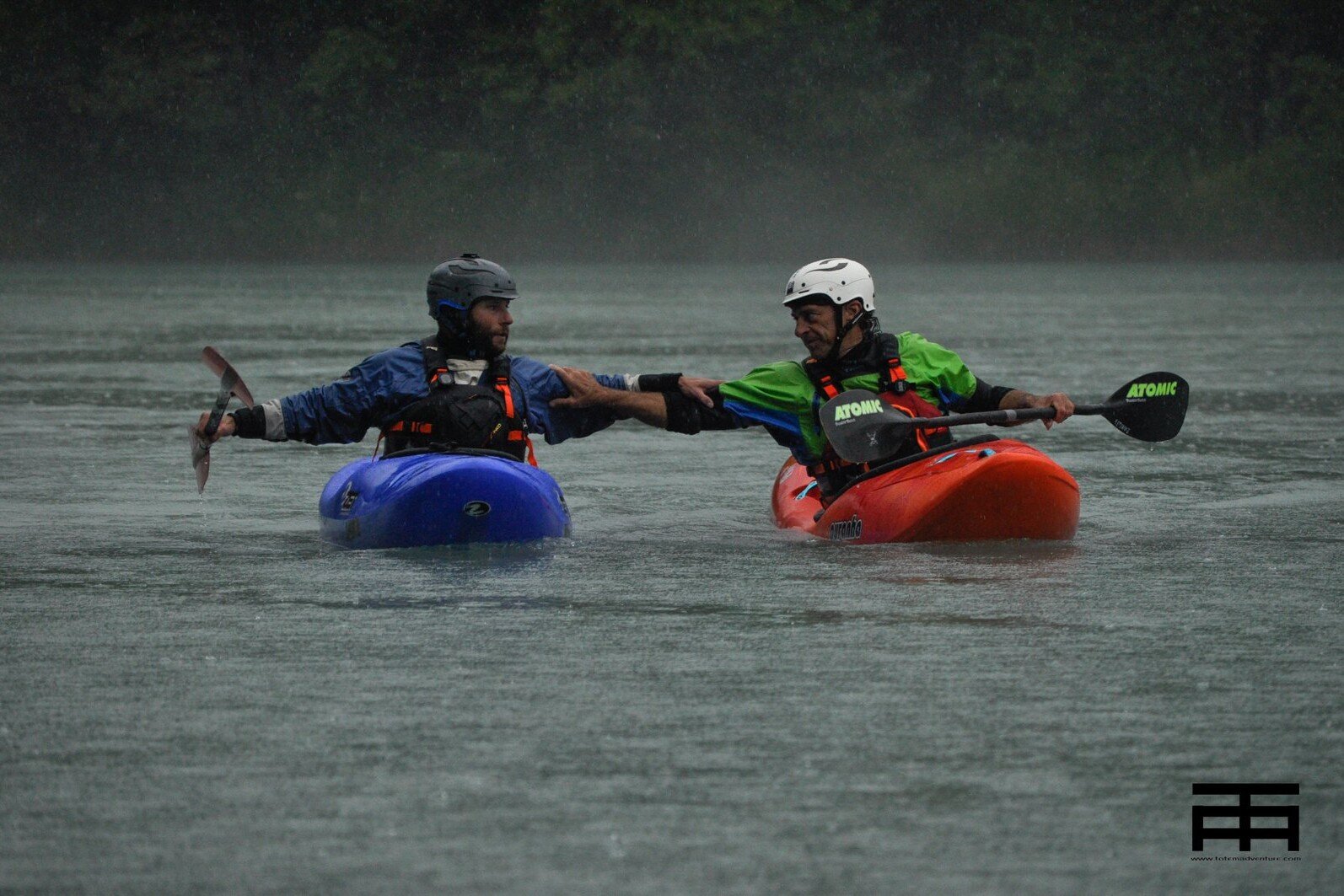 Totem Advenuture Two canoeists in blue and red kayaks shake hands on a rainy lake, sharing an exciting adventure that echoes the spirit of outdoor experiences. Rafting Kayaking Paragliding Canyoning Aosta Valley