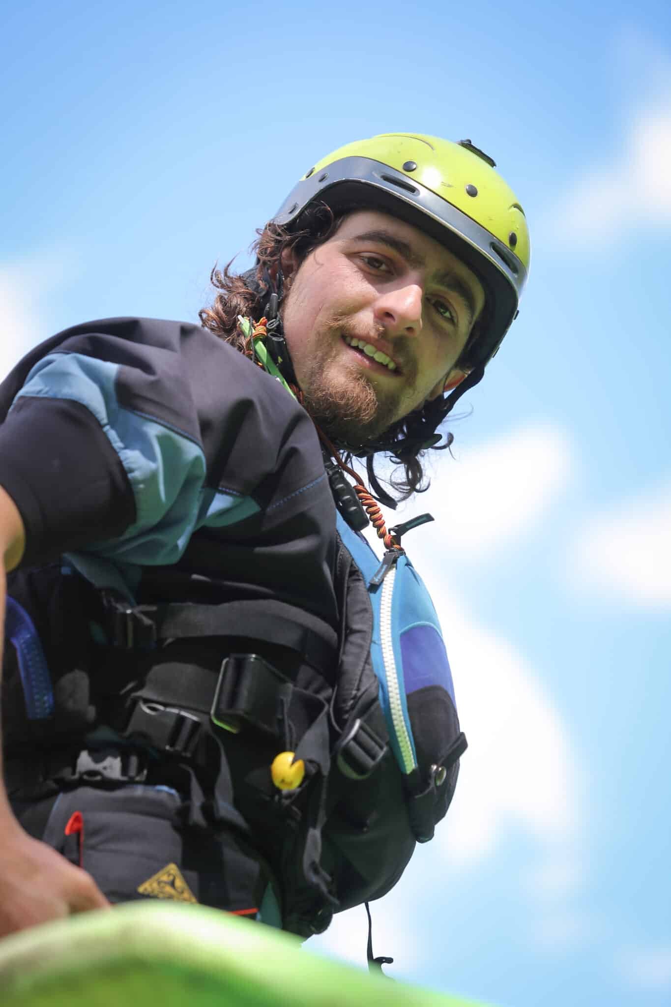 Totem Advenuture Un uomo con casco e attrezzatura da outdoor sorride, pronto per un'avventura del Team Totem, in piedi contro un cielo blu brillante con nuvole. Rafting Kayaking Parapendio Canyoning Valle d’Aosta