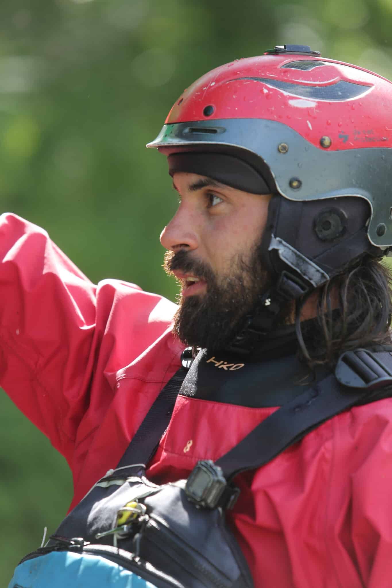 Totem Advenuture Un uomo con la barba che indossa un casco e una giacca rossa, forse in kayak con il Team Totem Adventure, alza il braccio all'aperto. Rafting Kayaking Parapendio Canyoning Valle d’Aosta
