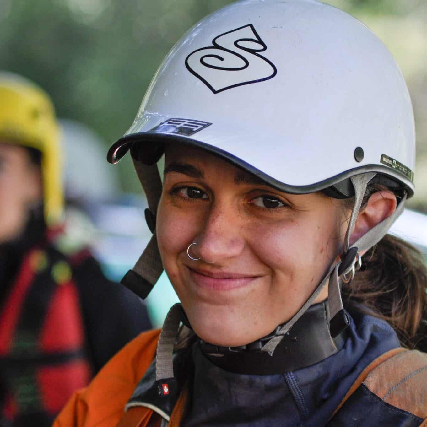 Totem Advenuture A woman with helmet and equipment smiles outdoors during a Team Totem adventure, with another person out of focus in the background. Rafting Kayaking Paragliding Canyoning Aosta Valley