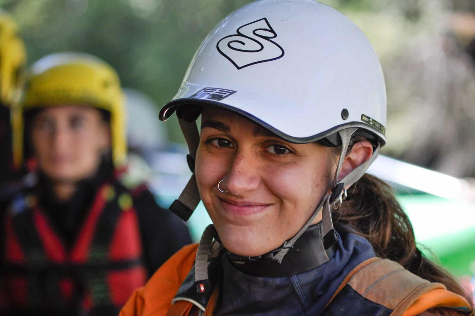 Totem Advenuture Una donna con casco e attrezzatura sorride all'aperto durante un'avventura del Team Totem, con un'altra persona sfocata sullo sfondo. Rafting Kayaking Parapendio Canyoning Valle d’Aosta