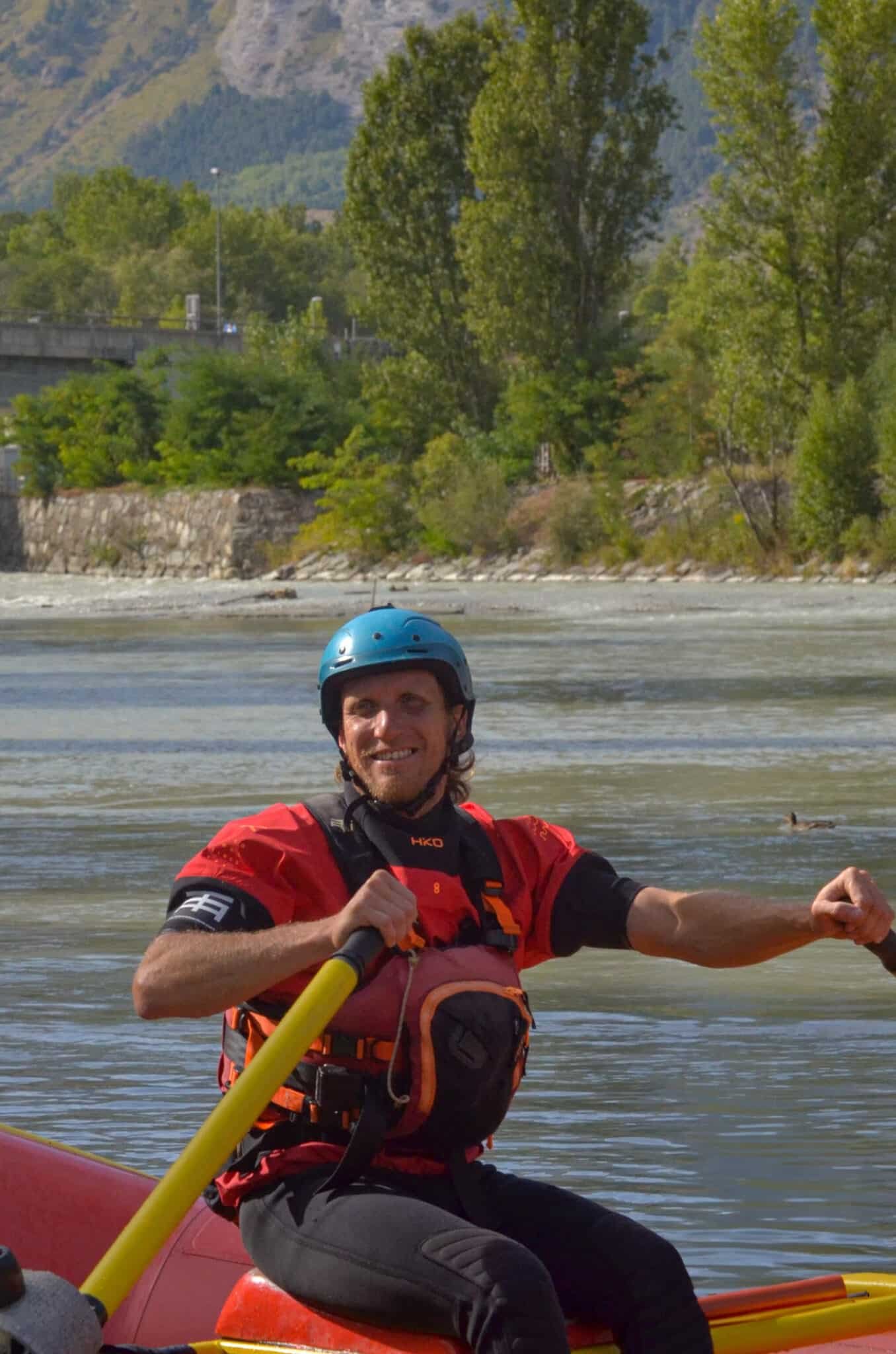 Totem Advenuture Uomo con casco e giubbotto di salvataggio che pagaia su una zattera rossa su un fiume, circondato da alberi e montagne, vivendo un'emozionante avventura del Team Totem. Rafting Kayaking Parapendio Canyoning Valle d’Aosta