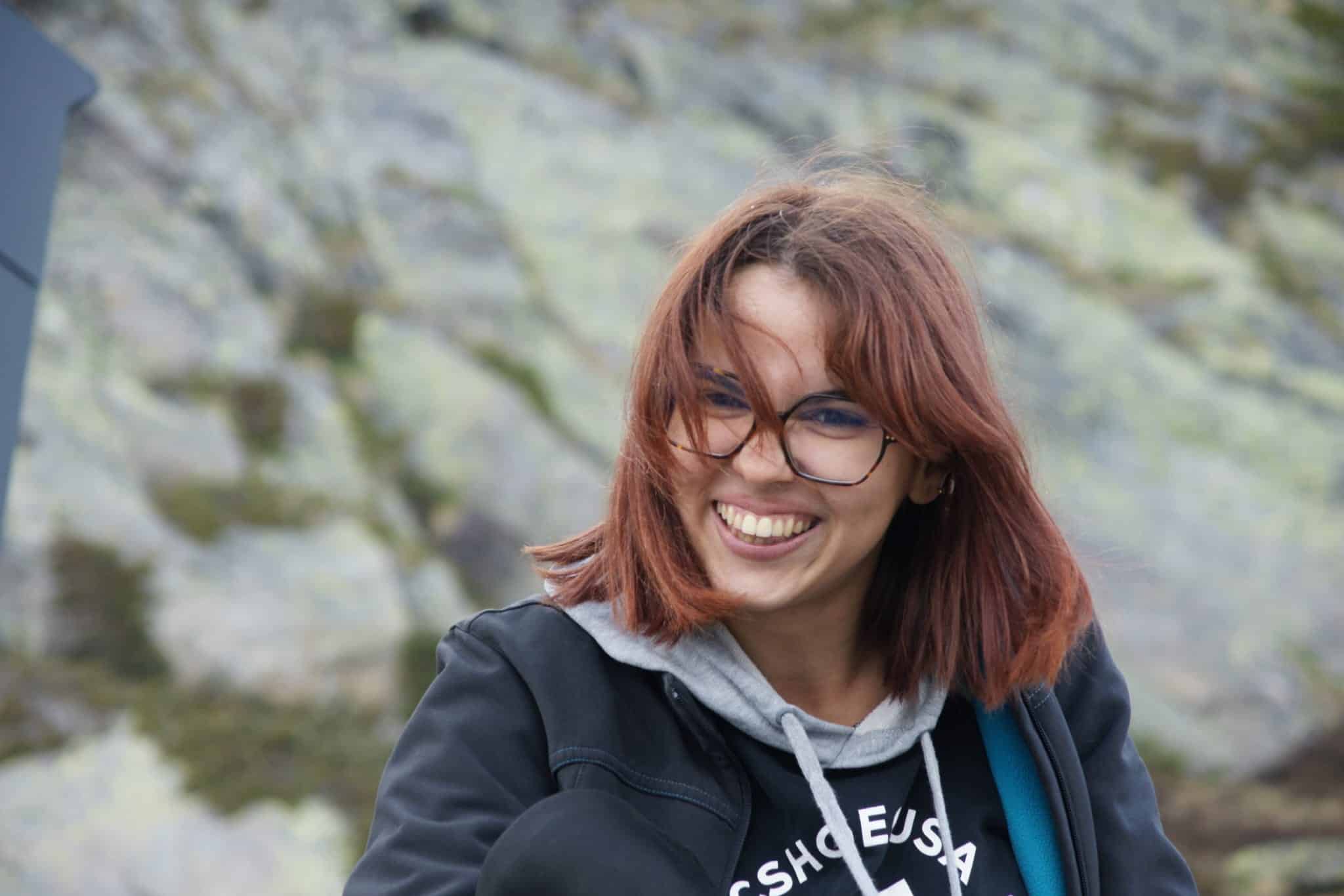 Totem Advenuture Una giovane donna con gli occhiali sorride all'aperto, con i capelli mossi dal vento, mentre si gode l'avventura di un totem di squadra su uno sfondo roccioso. Rafting Kayaking Parapendio Canyoning Valle d’Aosta