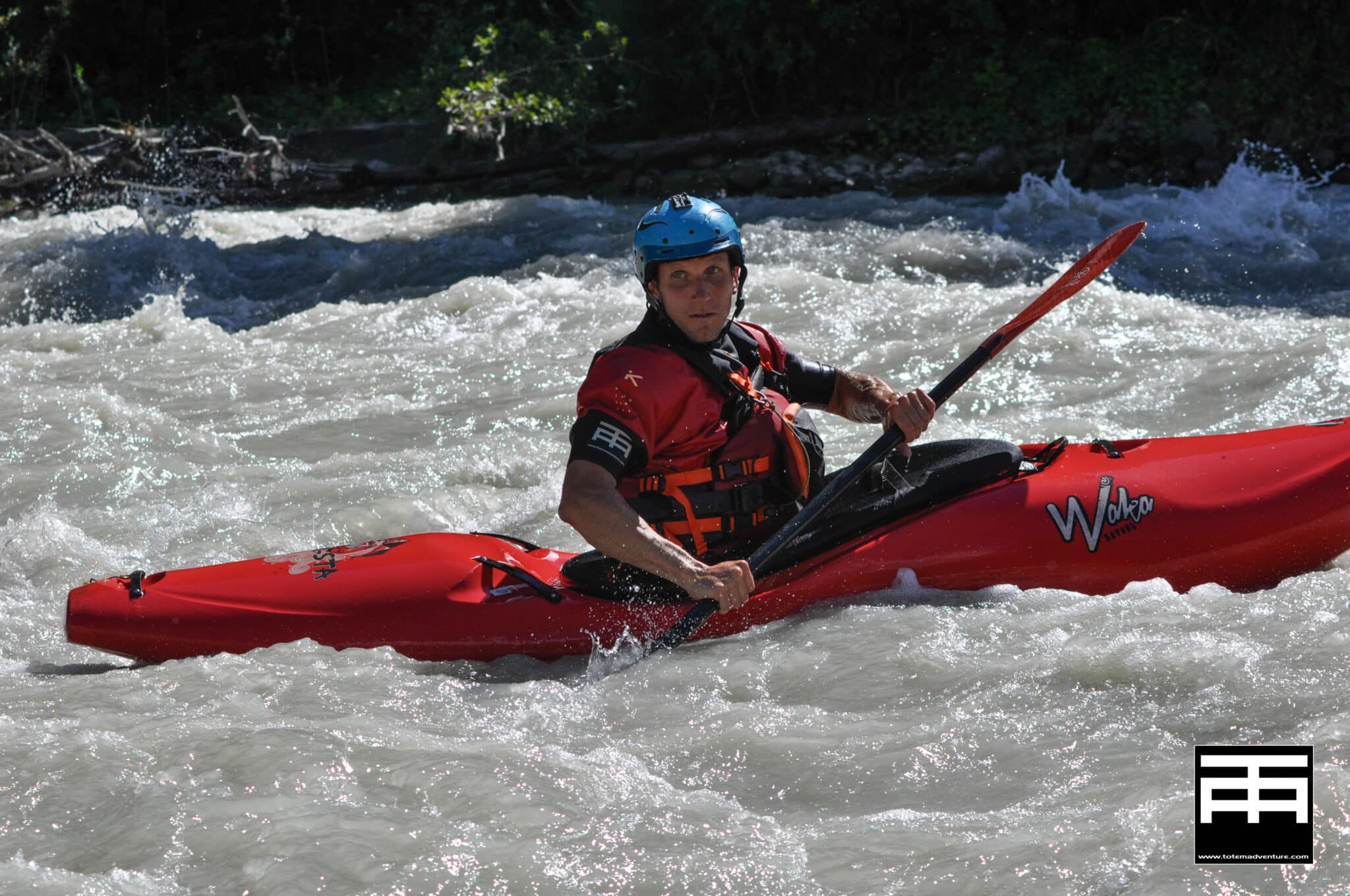 Totem Advenuture Una persona in un kayak rosso pagaia attraverso rapide d'acqua bianca in rapido movimento, indossando un casco e un giubbotto di salvataggio, abbracciando il brivido di un'avventura del Team Totem. Rafting Kayaking Parapendio Canyoning Valle d’Aosta