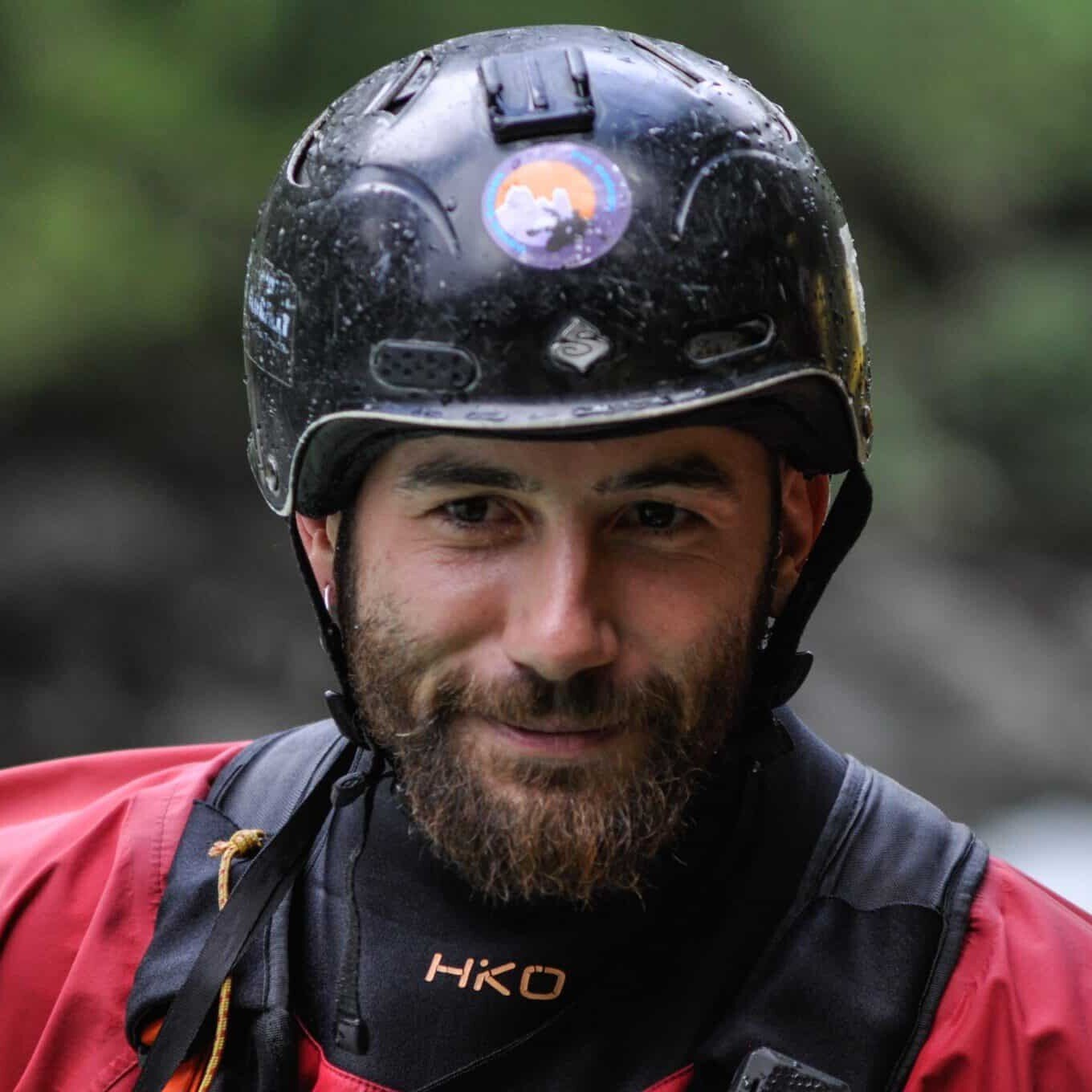 Totem Advenuture Un uomo in giacca rossa e casco nero sorride all'aperto, con uno sfondo naturale sfocato. Rafting Kayaking Parapendio Canyoning Valle d’Aosta