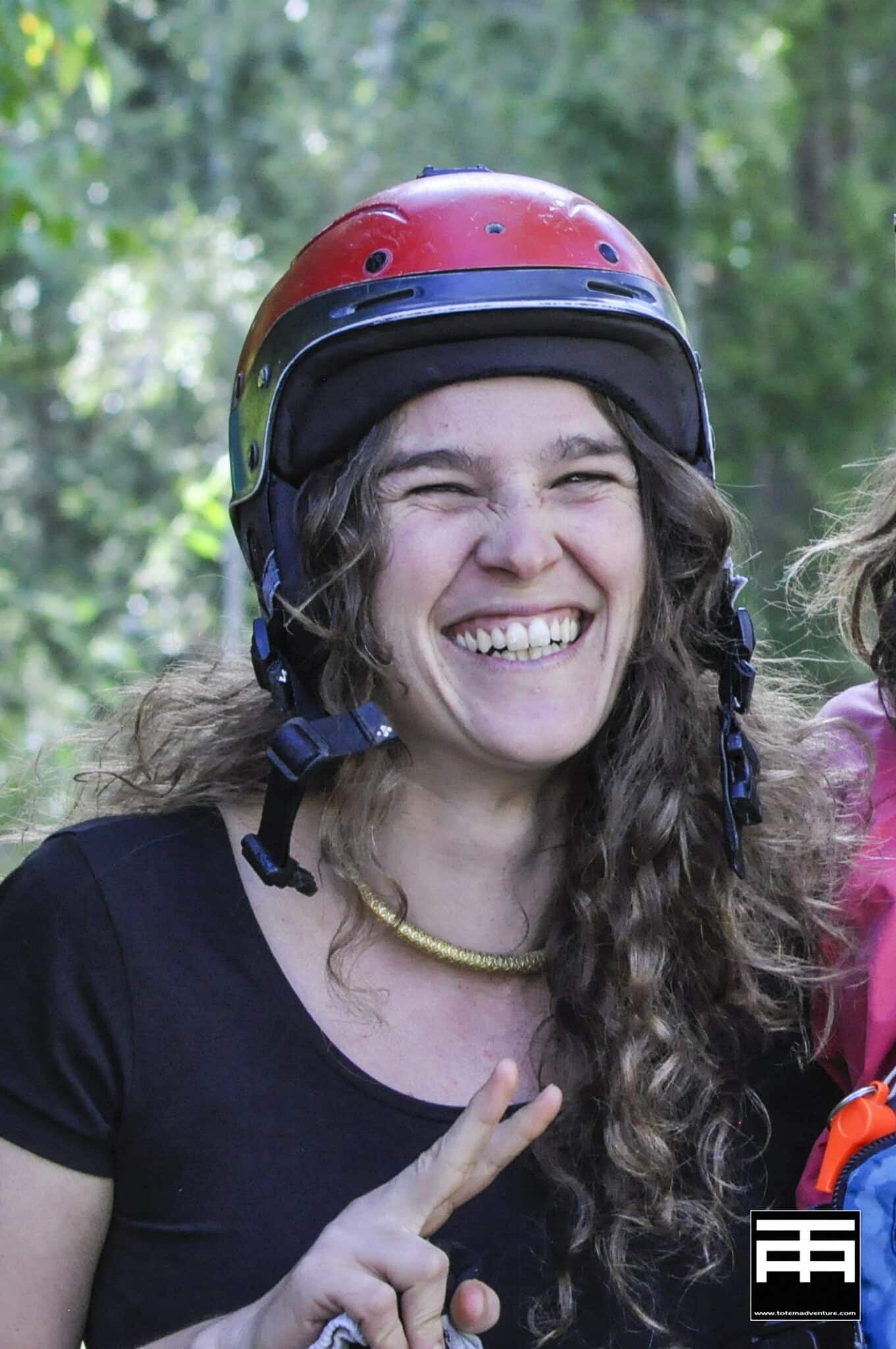 Totem Advenuture Una donna sorridente con un casco rosso fa il segno della pace, in piedi all'aperto con alberi verdi sullo sfondo, pronta per la prossima avventura del Team Totem. Rafting Kayaking Parapendio Canyoning Valle d’Aosta
