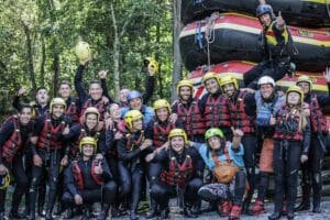 Totem Advenuture Un gruppo di avventurosi rafters con caschi e mute posa allegramente in mezzo alla foresta, con zattere accatastate dietro di loro, mostrando la loro emozionante esperienza all'aria aperta in Valle d'Aosta. Rafting Kayaking Parapendio Canyoning Valle d’Aosta