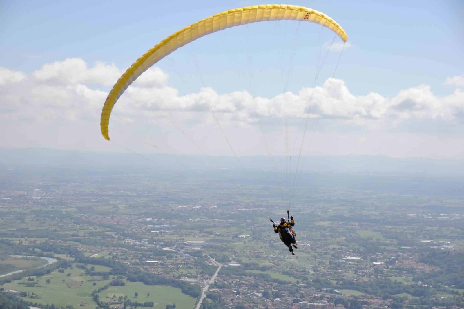 Totem Advenuture Due persone stanno volando in parapendio con un paracadute giallo sopra lo spettacolare paesaggio di Pré-Saint-Didier, dove campi e edifici sparsi si estendono più in basso, offrendo viste emozionanti quanto il rafting in Valle d'Aosta. Rafting Kayaking Parapendio Canyoning Valle d’Aosta