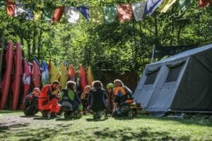 Totem Advenuture Un gruppo di persone in tenuta da esterno siede in cerchio vicino a kayak e una tenda, pronto per un'avventura sotto bandiere colorate. È l'ambiente perfetto per vivere la bellezza mozzafiato della Valle d'Aosta. Rafting Kayaking Parapendio Canyoning Valle d’Aosta