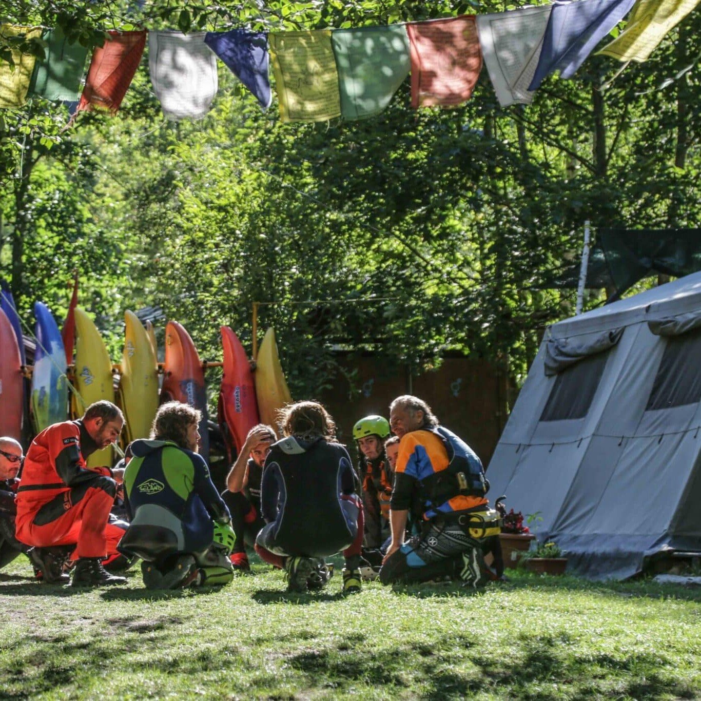 Totem Advenuture Un gruppo di persone in tenuta da esterno siede in cerchio vicino a kayak e una tenda, pronto per un'avventura sotto bandiere colorate. È l'ambiente perfetto per vivere la bellezza mozzafiato della Valle d'Aosta. Rafting Kayaking Parapendio Canyoning Valle d’Aosta