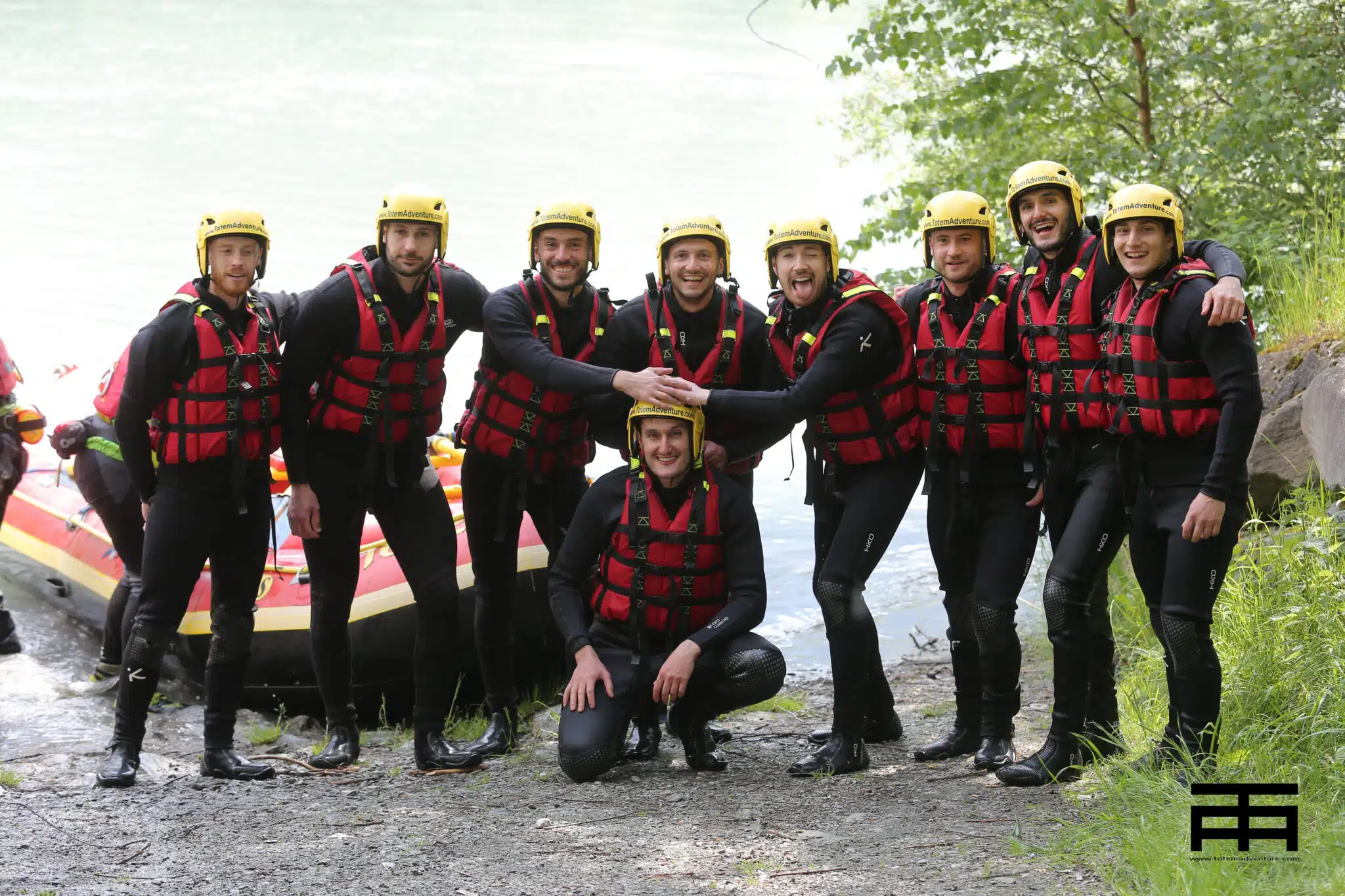 Totem Advenuture Un gruppo di persone con caschi e giubbotti di salvataggio posa davanti a un gommone in riva a un fiume, pronte a lanciarsi in un'entusiasmante avventura di rafting nella pittoresca Valle d'Aosta. Rafting Kayaking Parapendio Canyoning Valle d’Aosta