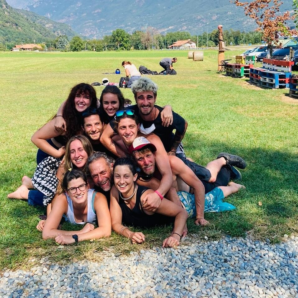 Totem Advenuture A group of ten smiling people, lying on a playful and adventurous totem pole on green grass, with mountains in the background. Rafting Kayaking Paragliding Canyoning Aosta Valley