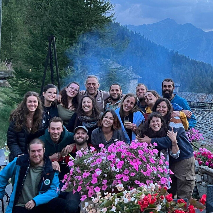 Totem Advenuture A group of people smiling outdoors, enjoying an adventure with a totem pole, with flowers and mountains in the background. Rafting Kayaking Paragliding Canyoning Aosta Valley
