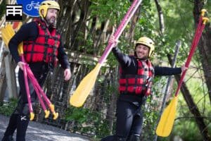 Totem Advenuture Due uomini sorridenti con casco e giubbotto di salvataggio trasportano pagaie gialle, preparandosi per un'avventura di rafting all'aperto. La loro eccitazione dimostra che per loro il rafting è normale, senza paura di fare rafting. Rafting Kayaking Parapendio Canyoning Valle d’Aosta
