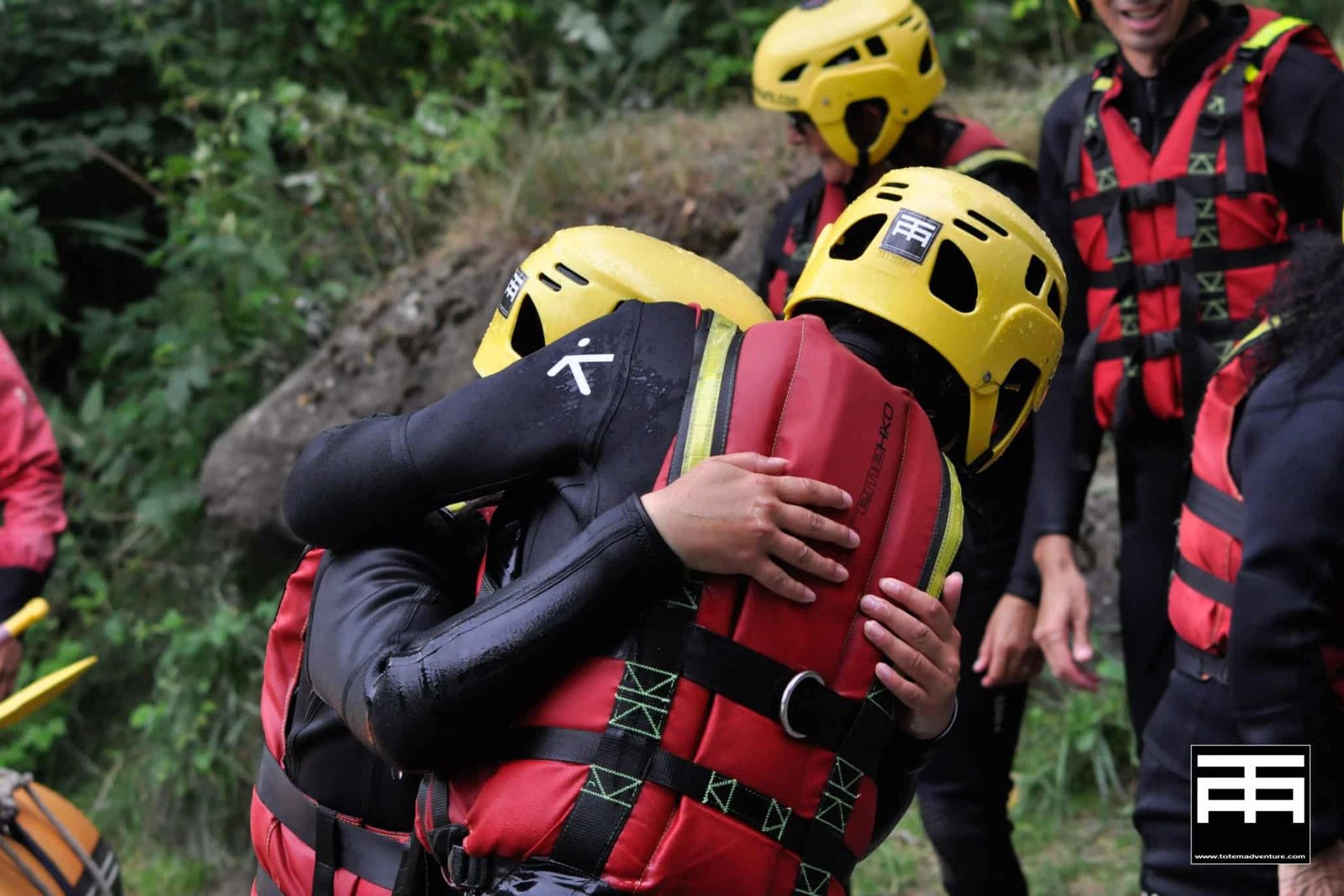 Totem Advenuture Due persone con casco e giubbotto di salvataggio si abbracciano all'aperto, circondate da altre persone in equipaggiamento di sicurezza, pronte per la loro prossima avventura con Bozza automatica. Rafting Kayaking Parapendio Canyoning Valle d’Aosta
