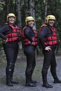 Totem Advenuture Tre persone con il casco, la muta e il giubbotto di salvataggio rosso sono in piedi all’aperto e sorridono davanti ad alcuni alberi. Rafting Kayaking Parapendio Canyoning Valle d’Aosta