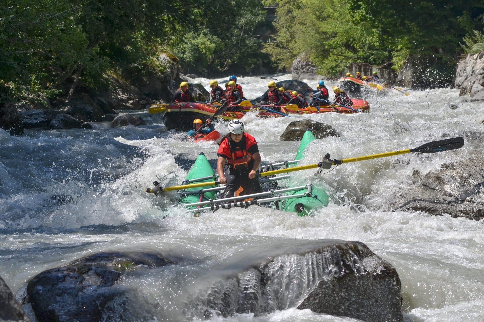 Totem Advenuture Persone con caschi e giubbotti di salvataggio affrontano le rapide su gommoni gonfiabili, circondati da rocce. Rafting Kayaking Parapendio Canyoning Valle d’Aosta